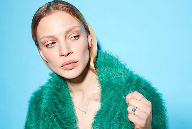Woman wearing a delicate white gold necklace and diamond ring with a blue center stone, styled with a teal faux-fur coat.