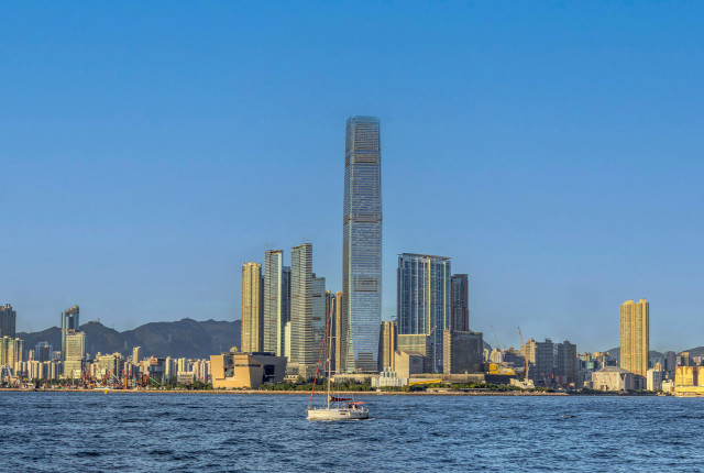 香港維港海景與天際線，前景白色帆船航行，中央高聳玻璃摩天大樓倒映藍天，呈現城市地標風景