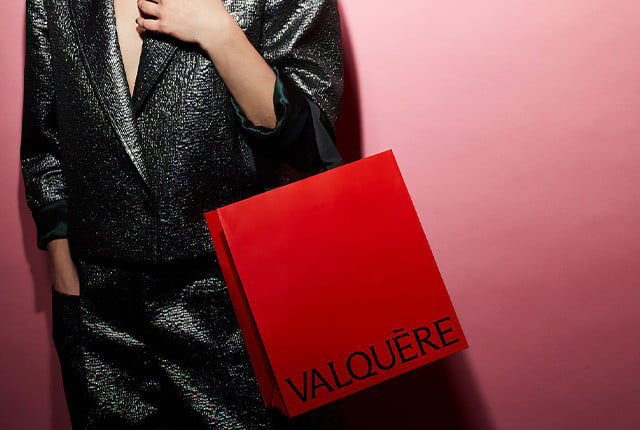 Shopping bag rosso lucido Valquère con logo nero, accanto a outfit elegante scuro scintillante